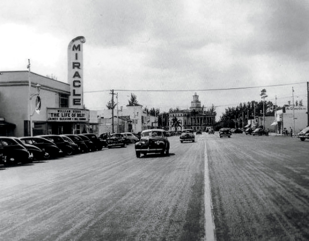 Black and white street view of Miracle Mile in Coral Gables during the 1940s, showing vintage cars, the Miracle Theatre marquee, and City Hall in the distance.