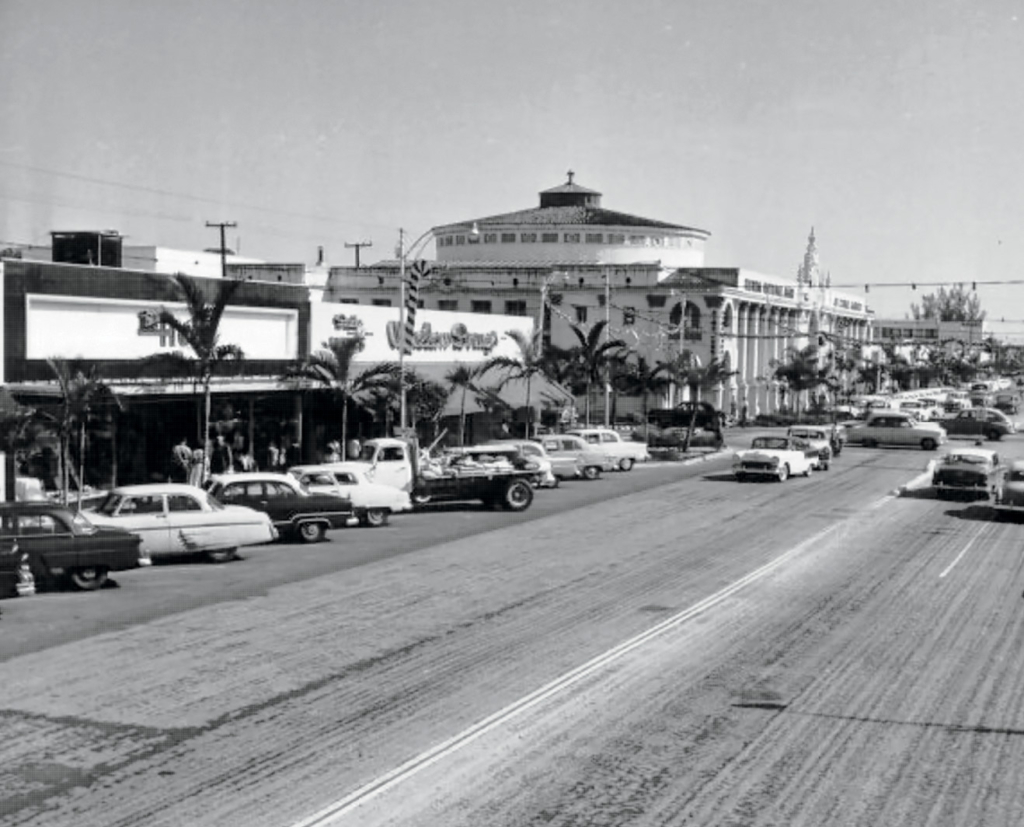 Street view of Miracle Mile in the 1960s showing vintage cars, bustling retail storefronts, palm trees, and the iconic Colonnade Building in Coral Gables.