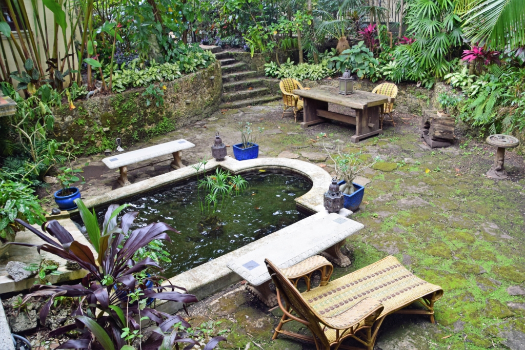 Historic Coral Gables sunken garden with koi pond, stone benches, tropical foliage, and rustic dining area in George Merrick-built home